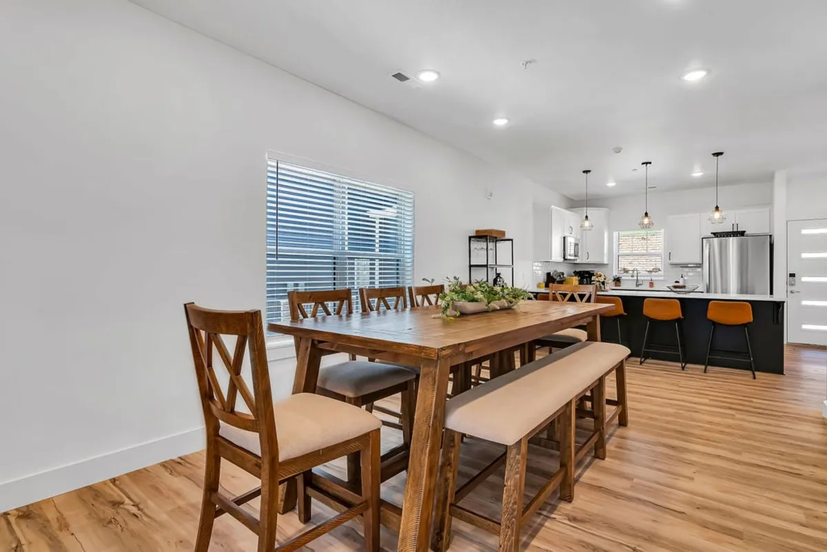 A welcoming dining area centered around a long wooden table, with additional bar stools at the kitchen island and a window letting in natural light. The space is set up for family meals, celebrations, and shared stories.