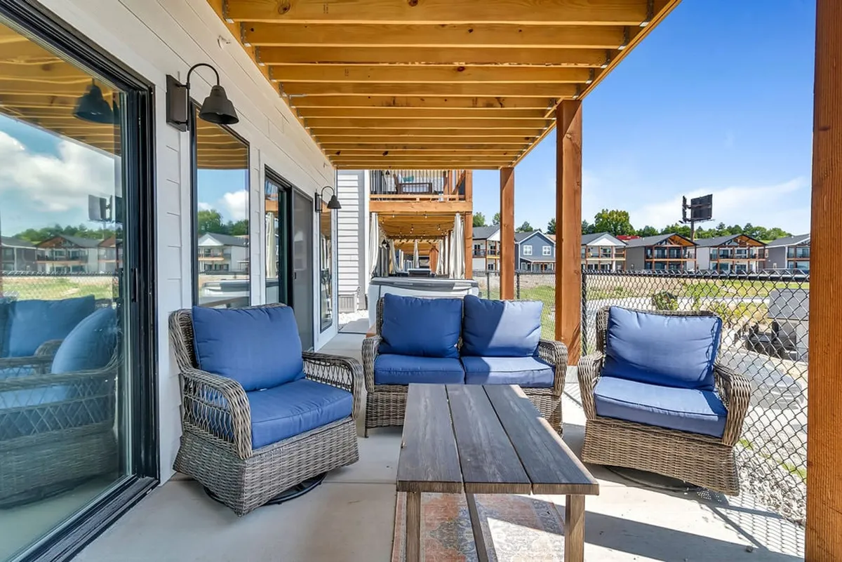 An exterior scene showcasing an outdoor seating area under sturdy wooden beams, with decorative lighting that creates a welcoming ambiance. The space is designed for socializing, enjoying shared meals, and taking in sunset or lake views from the deck.