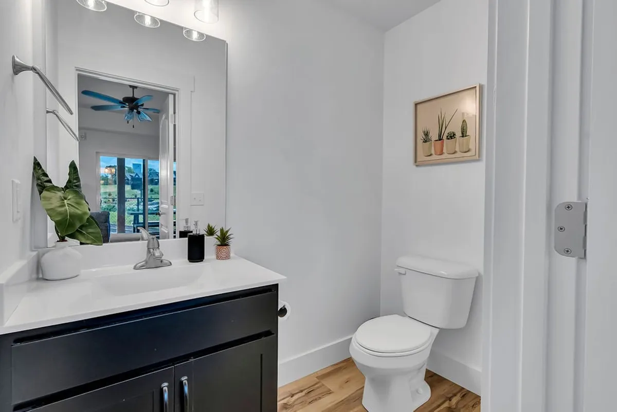 A bright, modern bathroom with a sleek sink, large mirror, small potted plants, toilet, and warm wooden flooring. The design is clean and inviting, embodying the relaxation and privacy of a high-end ensuite.