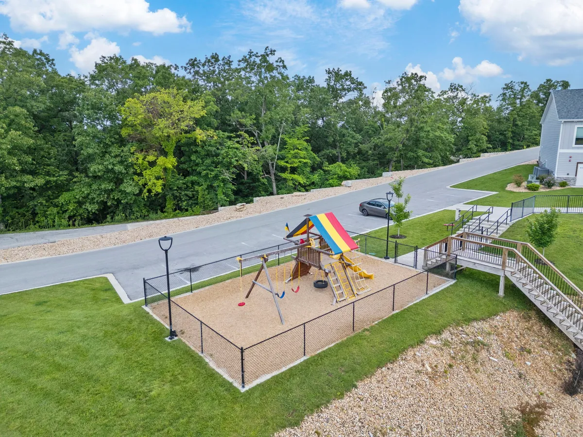 A vibrant children’s play area is shown, featuring a colorful play structure, swing set, and lush green grass surrounded by trees. Children are actively playing, while families watch and enjoy the space together, emphasizing a joyful, family-friendly environment.