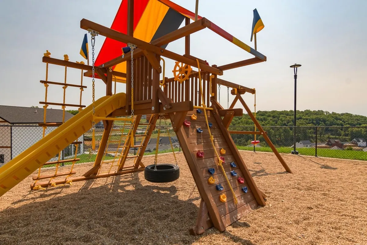 A cheerful outdoor playground scene featuring a colorful play structure, climbing wall, slide, tyre swing, and festive flags. Children are seen playing, climbing, and swinging, embodying a spirit of joyful, active recreation.