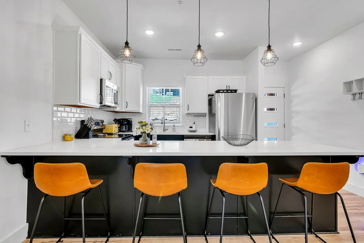 A modern, fully equipped kitchen with a bright kitchen island, orange stools, sleek appliances, and sunlight pouring in from the window. The open design invites both cooking and socializing.