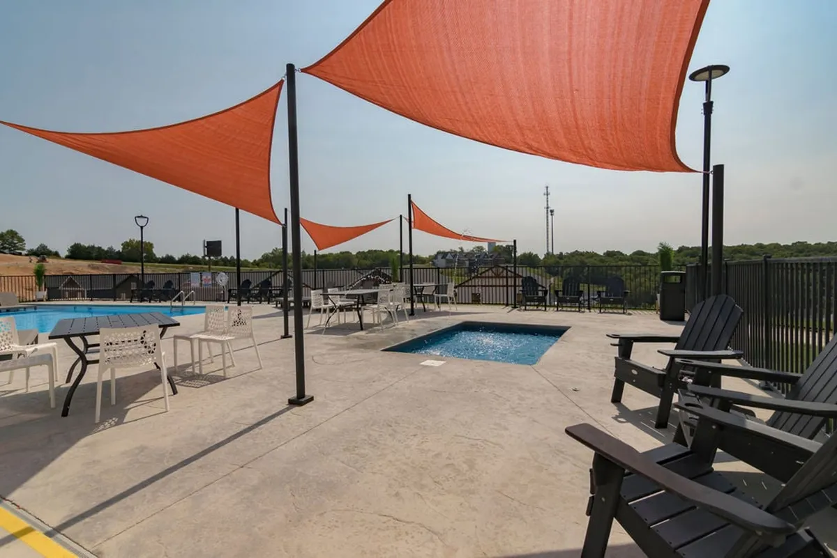 This exterior image captures a swimming pool with orange shade sails, outdoor seating areas, and a scenic landscape in the background. The cheerful atmosphere suggests opportunities for socializing, sunbathing, and enjoying the outdoors.