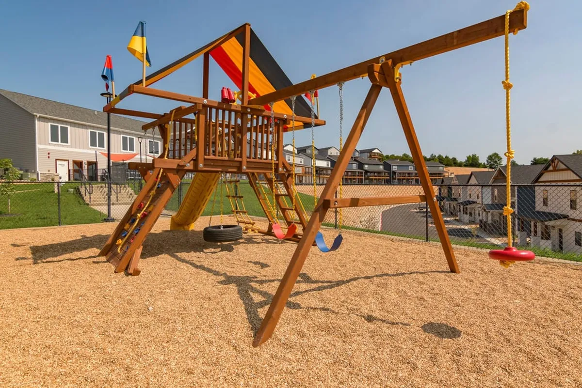 This image showcases a vibrant playground structure complete with swings, a slide, and colorful flags. Children are seen playing and families are gathered nearby, capturing the essence of outdoor fun and togetherness.