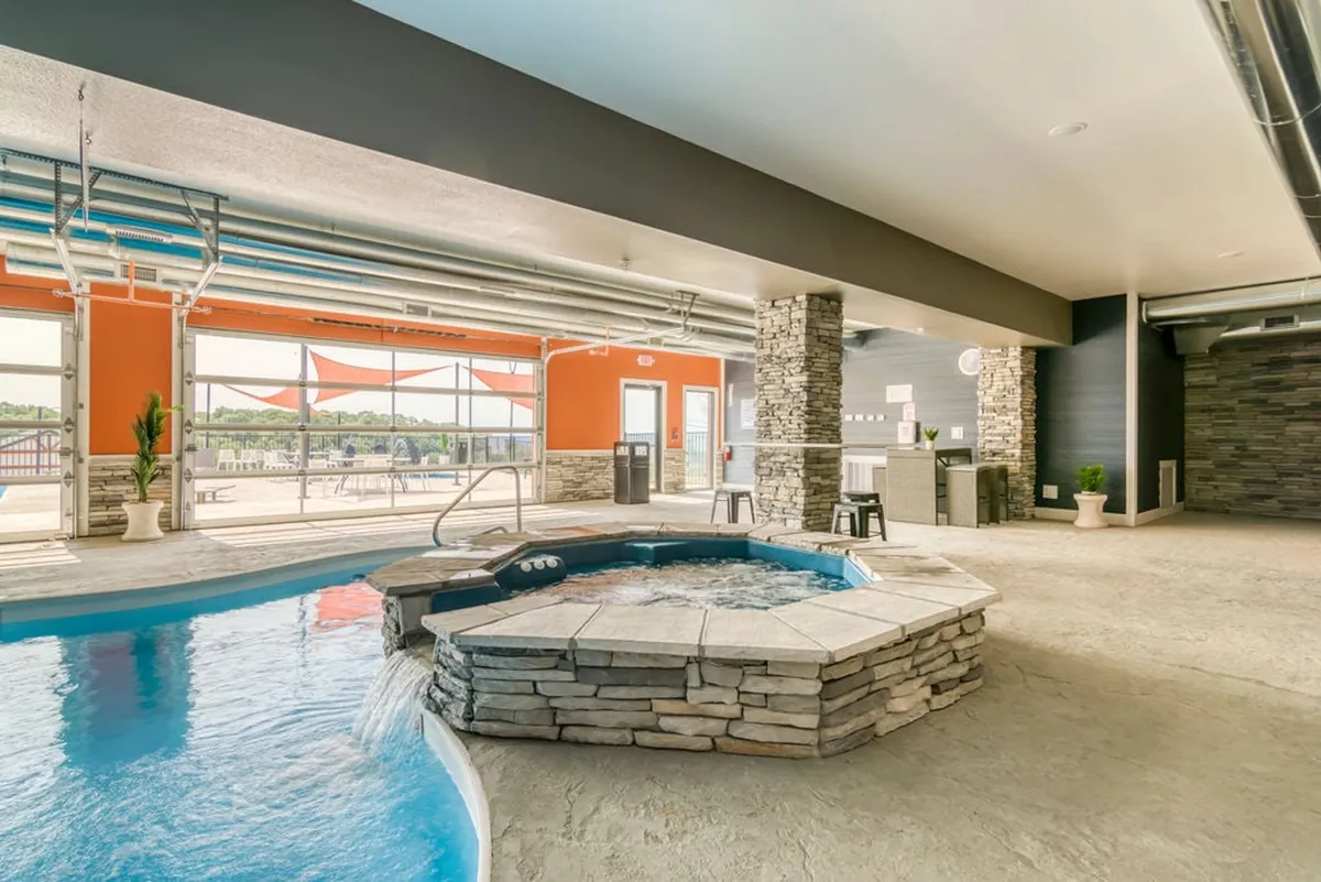 An interior view showcasing an inviting indoor pool and hot tub area, with large windows letting in natural light and a tranquil stone wall adding a touch of luxury. The space suggests year-round swimming and social relaxation regardless of the weather.