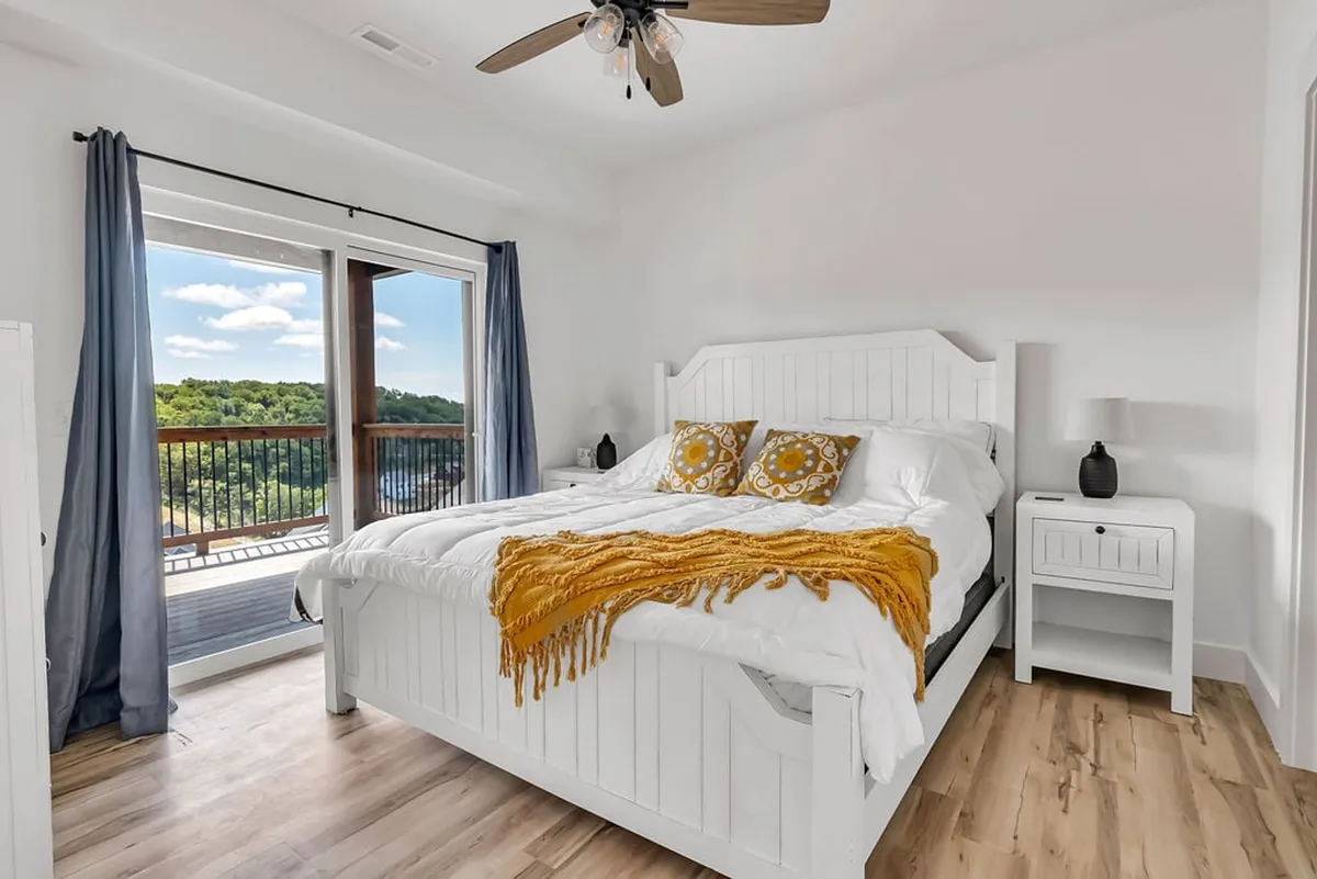 This image reveals a serene bedroom with a large, inviting bed adorned with decorative pillows and a soft yellow throw blanket. Expansive windows bathe the room in natural light, offering tranquil views and a cozy, restful atmosphere.
