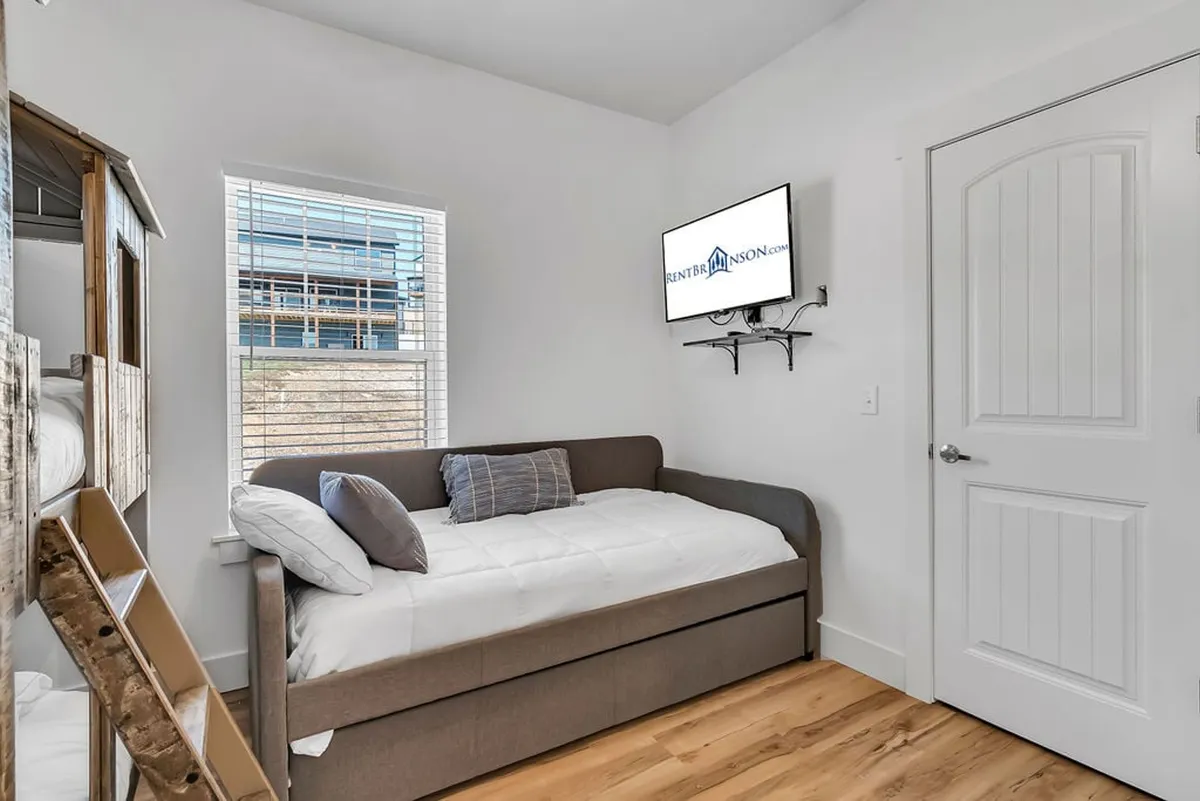 A playful and versatile bedroom featuring a daybed, TV, a window letting in natural light, decorative pillows, and a charming wooden playhouse. The space feels inviting—ideal for both relaxation and imaginative play.