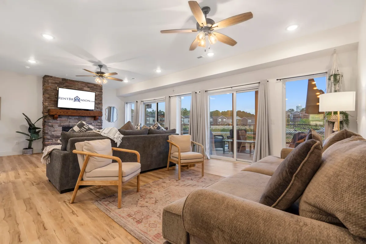The image shows a warm and inviting living area with plush seating arranged around a striking stone feature wall, large windows that fill the room with natural light, and a cozy atmosphere ideal for relaxing or gathering with loved ones.
